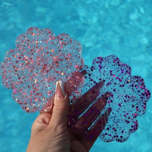 May include: Two decorative resin coasters with a floral shape. One coaster is pink with pink glitter, and the other is blue with purple glitter. The coasters are held against a blue water background.