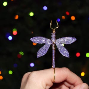 May include: A purple glitter dragonfly ornament with gold wire accents. The dragonfly has outstretched wings and is held by a gold ring. The background is a dark green with colorful bokeh lights.