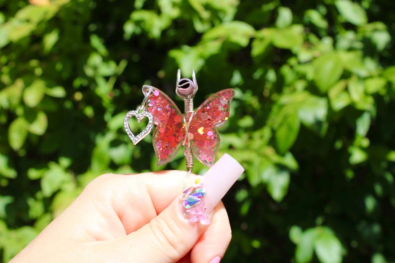 Image shows a cigarette holder that is a ring that has a wire wrapped pink sequin butterfly with a rhinestone heart charm. The top has prongs that is adjustable wire.