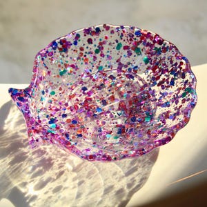 May include: A clear, decorative bowl with a scalloped edge, filled with colorful glitter. The glitter includes red, blue, purple, and green specks. The bowl casts a shadow on a white surface.