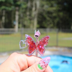Image shows a cigarette holder that is a ring that has a wire wrapped pink sequin butterfly with a rhinestone heart charm. The top has prongs that is adjustable wire.
