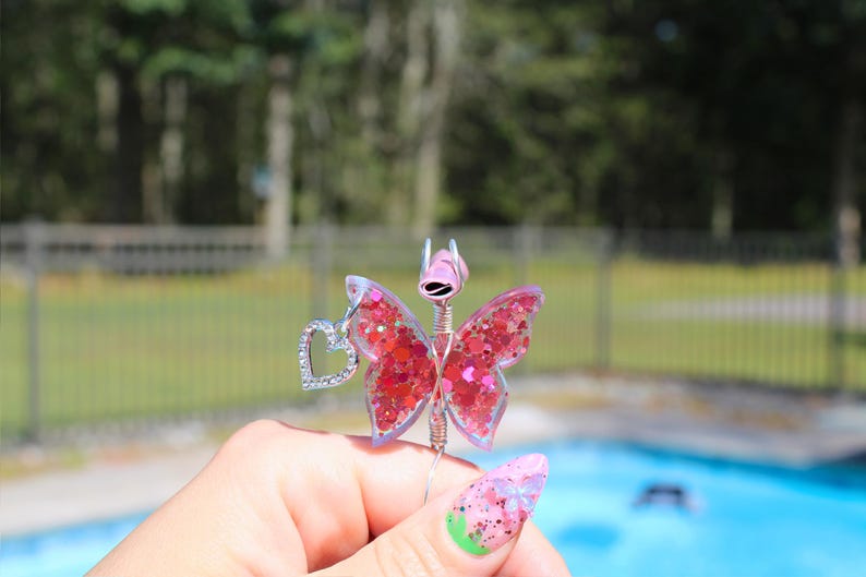 Image shows a cigarette holder that is a ring that has a wire wrapped pink sequin butterfly with a rhinestone heart charm. The top has prongs that is adjustable wire.