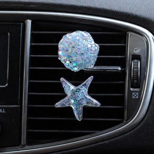May include: Two car air vent decorations. One is a round shape, the other is a starfish shape. Both are covered in iridescent glitter. The decorations are placed on a car's air vent.