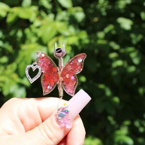 Image shows a cigarette holder that is a ring that has a wire wrapped pink sequin butterfly with a rhinestone heart charm. The top has prongs that is adjustable wire.