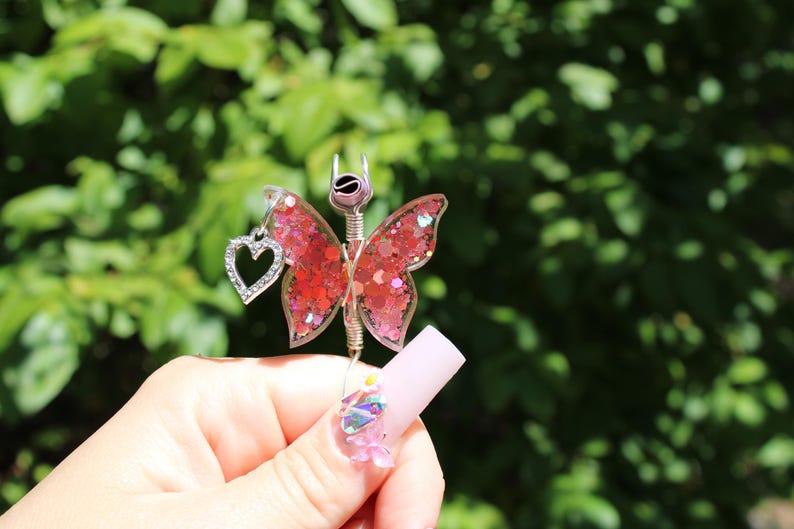 Image shows a cigarette holder that is a ring that has a wire wrapped pink sequin butterfly with a rhinestone heart charm. The top has prongs that is adjustable wire.