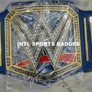 May include: A professional wrestling championship belt with a blue strap and gold-colored plates. The central plate features the WWE logo with the words "UNIVERSAL CHAMPION". The side plates also display the WWE logo.
