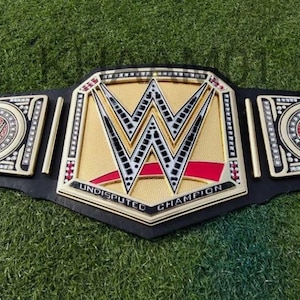 Puede incluir: Un cinturón de campeonato de lucha libre dorado y negro con el logotipo de la WWE. El cinturón presenta las palabras "UNDISPUTED CHAMPION" y está sobre un fondo verde.