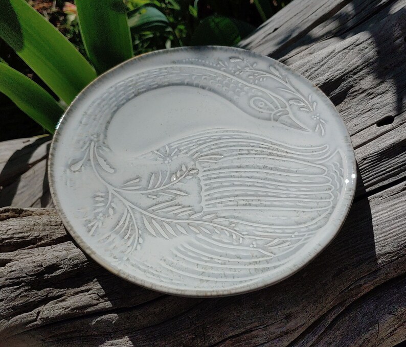 May include: A white ceramic plate featuring an embossed design of a bird with outstretched wings. The plate has a subtle grey glaze.