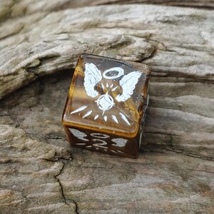 May include: A square, translucent, amber-colored stone with white angel wing and halo designs. The stone has a tiger's eye appearance and is set against a natural wood background. The design is repeated on multiple sides.