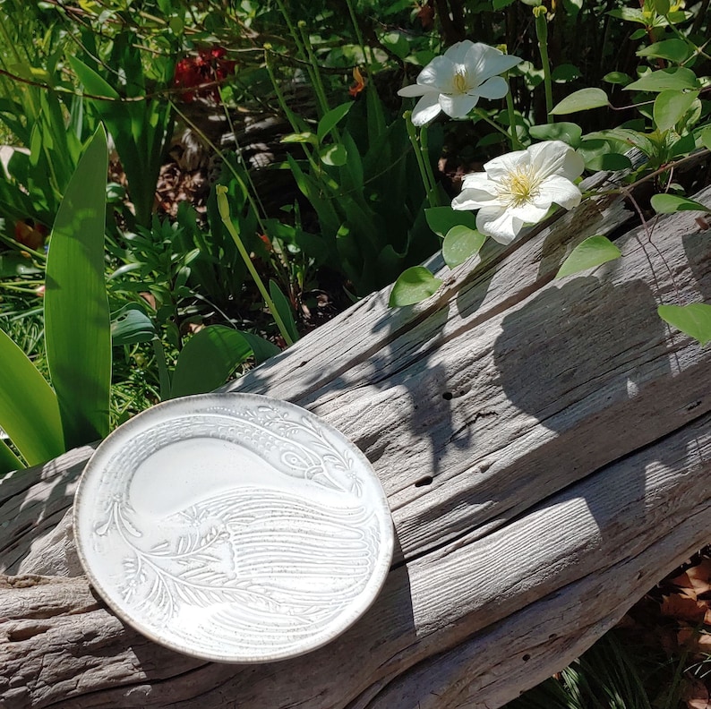 May include: A white ceramic plate with a raised design of a stylised bird. The plate is sitting on a weathered wooden surface.