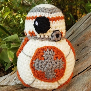 May include: A handmade crochet BB-8 droid. The spherical body is primarily white with orange and gray accents. The head features a black eye and gray details. The droid is made of yarn and sits on a piece of wood.