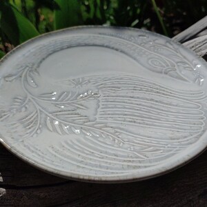 May include: A white ceramic plate featuring a raised design of a stylised bird with a long tail. The plate has a subtle speckled glaze.