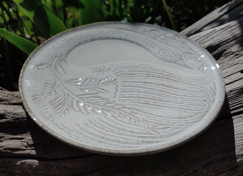 May include: A white ceramic plate featuring a raised design of a stylised bird with a long tail. The plate has a subtle speckled glaze.