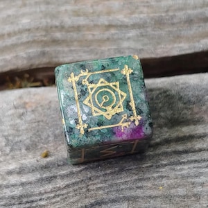 May include: A green and gold dice with a star-shaped symbol in the center. The dice is made of a natural stone with a speckled surface.