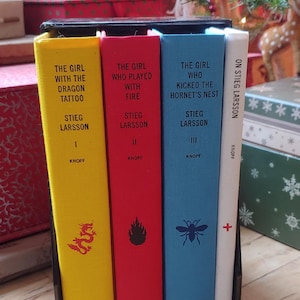 May include: A set of four books by Stieg Larsson, displayed in a black holder. The book spines are yellow, red, blue, and white, with titles including "The Girl with the Dragon Tattoo." The books are in front of a Christmas tree.