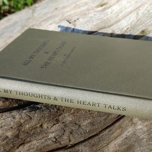 Puede incluir: Un libro de tapa dura titulado "ALL MY THOUGHTS & THE HEART TALKS" en un color verde apagado. El libro est&aacute; sobre una superficie de madera envejecida, con el texto del t&iacute;tulo visible en el lomo y la portada. Los bordes del libro est&aacute;n ligeramente desgastados.