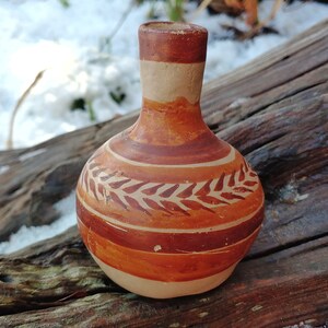 May include: A small, brown clay vase with a narrow neck and a wide, rounded body. The vase is decorated with a pattern of brown leaves on a white background.