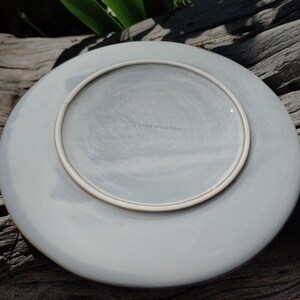 May include: A white ceramic plate with a light grey glaze and a brown rim. The plate has a subtle marbled effect and the text "Anthropologie" is printed on the bottom.