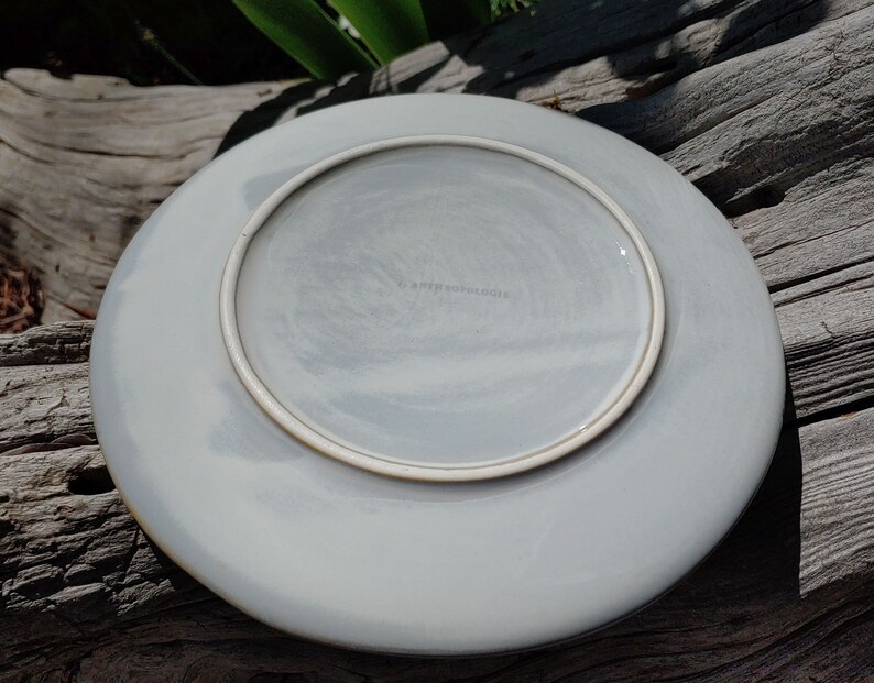 May include: A white ceramic plate with a light grey glaze and a brown rim. The plate has a subtle marbled effect and the text "Anthropologie" is printed on the bottom.