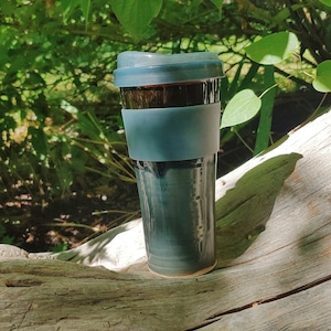 May include: A dark blue ceramic travel mug with a silicone lid and a blue silicone band around the middle. The mug is sitting on a piece of driftwood.