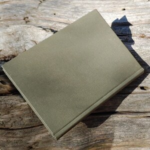 Puede incluir: Un libro cerrado, de color verde oliva, con una cubierta texturizada, descansa sobre una superficie de madera envejecida. La forma rectangular del libro y la sombra proyectada por la luz crean un contraste. La cubierta parece ser de un material duradero.