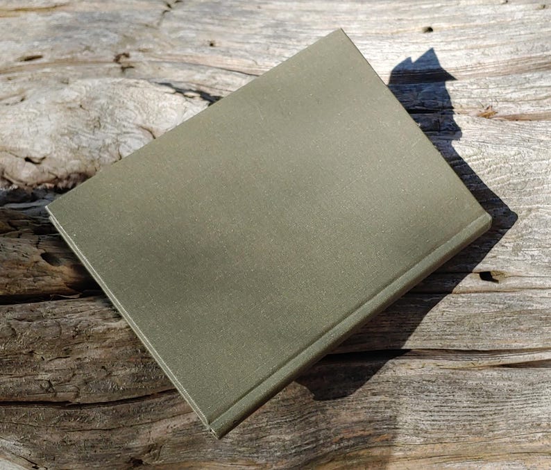 Puede incluir: Un libro cerrado, de color verde oliva, con una cubierta texturizada, descansa sobre una superficie de madera envejecida. La forma rectangular del libro y la sombra proyectada por la luz crean un contraste. La cubierta parece ser de un material duradero.