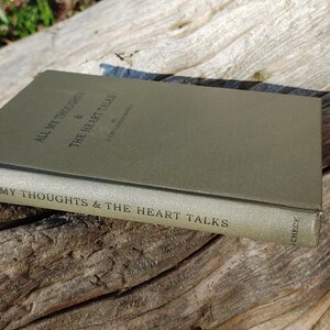 Puede incluir: Un libro verde oliva titulado "All My Thoughts & The Heart Talks" est&aacute; abierto sobre una superficie de madera envejecida. El lomo del libro muestra el t&iacute;tulo, y la cubierta es de un verde liso y apagado. La escena est&aacute; iluminada por luz natural.