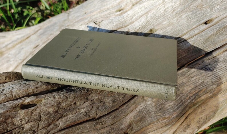 Puede incluir: Un libro verde oliva titulado "All My Thoughts & The Heart Talks" est&aacute; abierto sobre una superficie de madera envejecida. El lomo del libro muestra el t&iacute;tulo, y la cubierta es de un verde liso y apagado. La escena est&aacute; iluminada por luz natural.