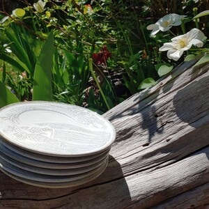 May include: A stack of five white ceramic plates with a decorative design. The plates are sitting on a weathered wooden log.