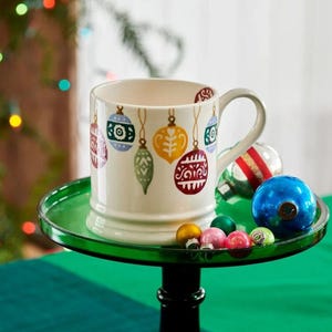 May include: A cream-colored ceramic mug decorated with colorful Christmas ornament designs. The mug sits on a green glass stand, surrounded by small, colorful ornaments. The mug has a handle and is filled with nothing.