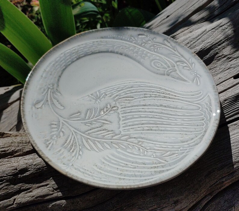 May include: White ceramic plate featuring an embossed design of a bird with outstretched wings. The design is in a light grey colour and has a subtle texture.