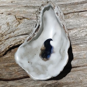 May include: A handmade ceramic oyster shell dish with a white interior and a dark blue and brown abstract design. The dish has a textured, off-white exterior and is displayed on a weathered wooden surface.