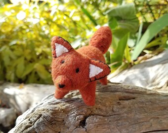 May include: A small, orange felt fox toy with black eyes and white felt ears. The fox is standing on a piece of weathered wood.