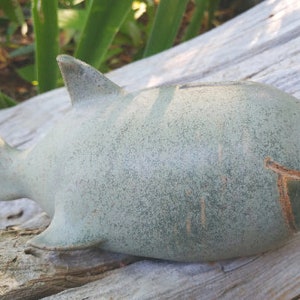 May include: A ceramic shark-shaped bank in a muted green hue. The shark has a dorsal fin, tail fin, and a slot on top for inserting coins. The mouth is open, revealing a carved detail. The bank is resting on a weathered wooden surface.
