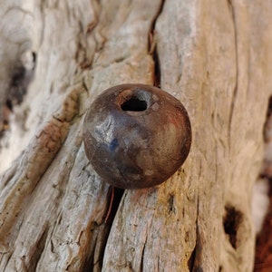May include: A dark brown, spherical bead with a hole through the center. The bead has a rough, textured surface and appears to be made of a natural material. The bead is displayed on a weathered, light-colored wooden surface.
