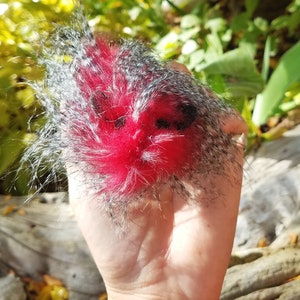 May include: A handmade, fluffy, red and gray decorative item. The central portion is a vibrant red, surrounded by a halo of gray and black fibers. The item is held in a hand, with a natural, blurred background of foliage.
