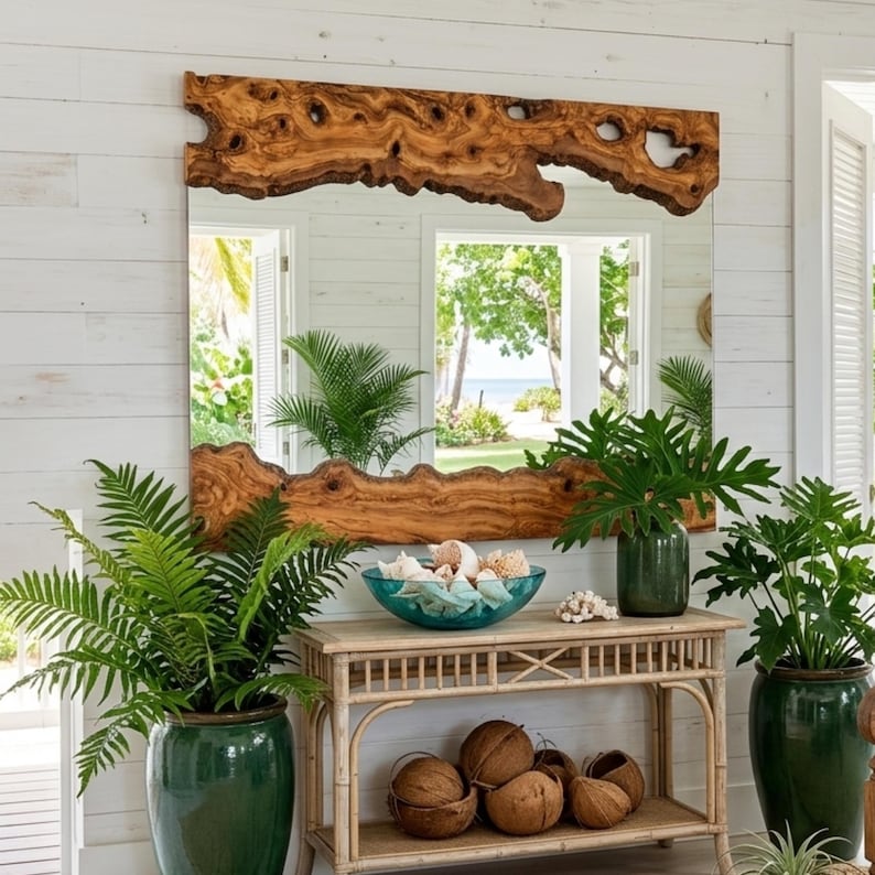 May include: A large rectangular mirror with a natural wood frame. The frame has a unique, organic shape with a light brown color. The mirror reflects a view of a beach and greenery. The mirror is mounted on a white wall.