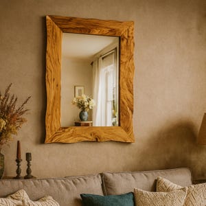 May include: A rectangular wooden mirror with a natural, textured frame hangs on a beige wall. The mirror reflects a room with a vase of flowers and a window with white curtains. The frame has a rustic, organic design.
