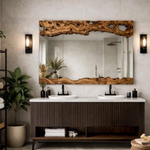 May include: A large rectangular mirror with a live edge wooden frame hangs above a double sink vanity. The vanity is dark brown with vertical grooves, and the countertop is white. Two black sconces flank the mirror.