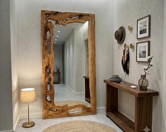 May include: A large floor mirror with a thick, natural-edged wooden frame featuring organic cutouts. The mirror is positioned in a hallway next to a wooden console table, a round jute rug, a table lamp, wall-mounted hooks, and framed art.