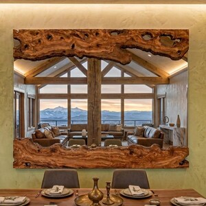 May include: Large rectangular mirror with a natural wood frame. The frame has a unique, organic shape and a warm, brown color. The mirror reflects a living room with a mountain view.