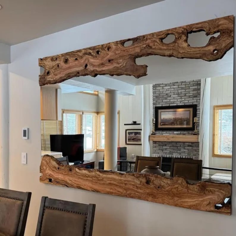 May include: A large rectangular mirror with a natural wood frame. The frame is made from a thick, irregularly shaped piece of wood with a light brown color and visible knots and holes. The mirror reflects a room with a fireplace and windows.