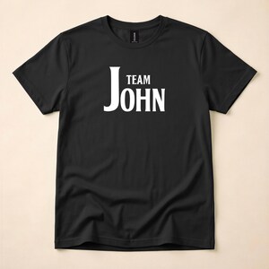 A black crewneck t-shirt featuring Team John in white text on the chest, laid flat on a neutral background.