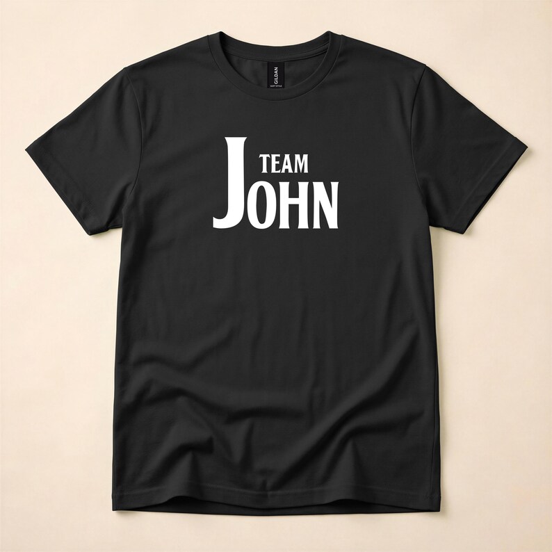 A black crewneck t-shirt featuring Team John in white text on the chest, laid flat on a neutral background.