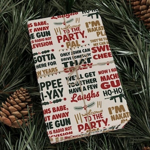 May include: A wrapped gift with a festive design. The wrapping paper is cream-colored and covered in phrases in red, green, and gold. The gift is tied with twine and rests on a bed of pine needles and pine cones.