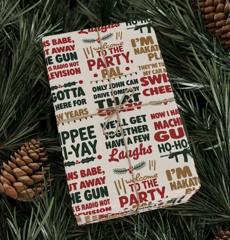 May include: A wrapped gift with a festive design. The wrapping paper is cream-colored and covered in phrases in red, green, and gold. The gift is tied with twine and rests on a bed of pine needles and pine cones.