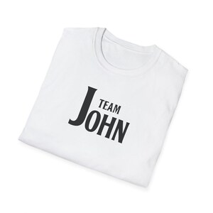 A folded white t-shirt angled on a white surface, featuring black text on the chest that reads Team John.