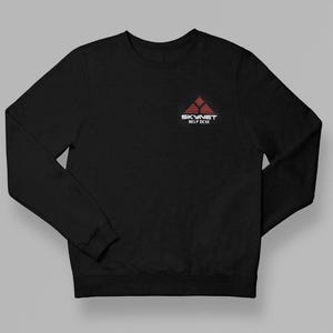 Könnte beinhalten: Ein schwarzes Langarm-Sweatshirt mit Rundhalsausschnitt und einer kleinen roten, dreieckigen Grafik auf der linken Brust. Die Grafik besteht aus horizontalen Linien und dem weißen Schriftzug SKYNET HELP DESK unter dem roten Dreieck.
