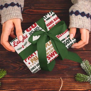 May include: A wrapped gift with a green ribbon is held by a person wearing a gray sweater. The wrapping paper has a white background with red and green text. The text includes phrases like "Laughs", "Ho-Ho", and "HANS BAB PUT AM".