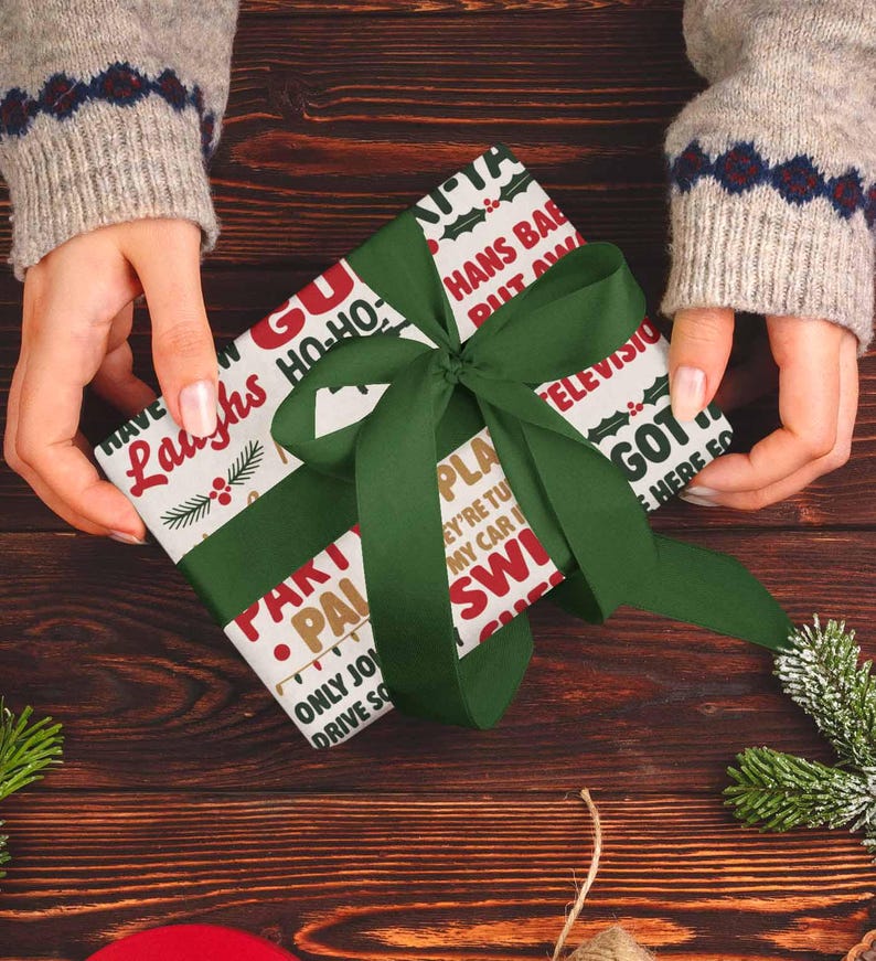 May include: A wrapped gift with a green ribbon is held by a person wearing a gray sweater. The wrapping paper has a white background with red and green text. The text includes phrases like "Laughs", "Ho-Ho", and "HANS BAB PUT AM".